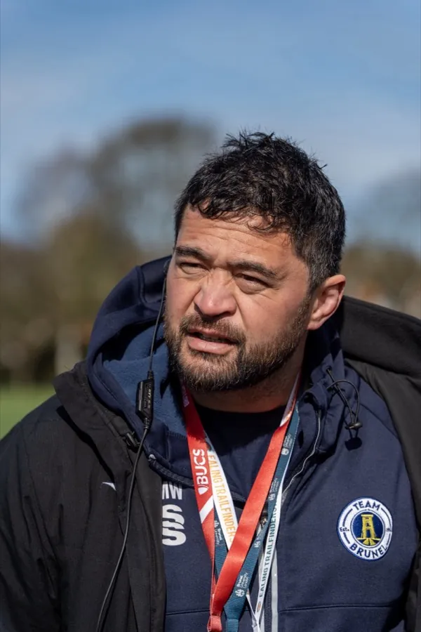 A member of the Team Brunel staff in a navy club jacket and BUCS lanyard watches from the touchline.