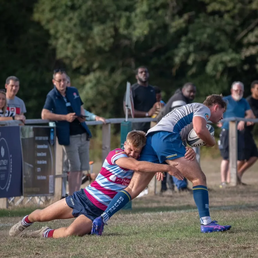 A Brunel ball carrier is brought down by a low tackle in front of the spectators lining the rail.