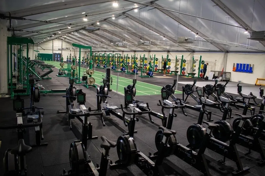 An indoor strength and conditioning hall with rows of rowing machines, weight racks and lifting platforms under a curved roof.