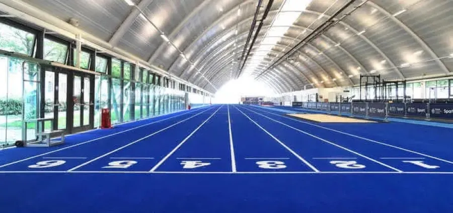 A blue six-lane indoor running track running the length of a bright curved-roof sports hall.