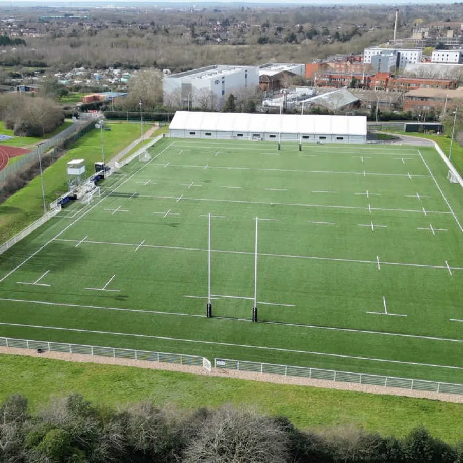 An aerial view of a full-size floodlit rugby pitch with posts and white markings, buildings and rooftops beyond.