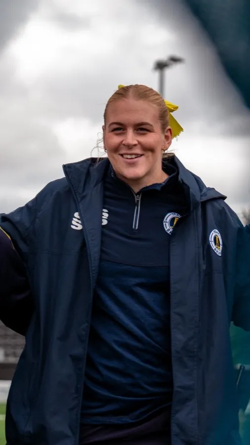 A member of the Team Brunel women's staff stands pitchside in a club coat and quarter-zip carrying the Team Brunel crest.