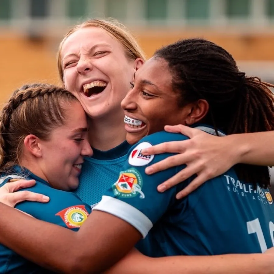 Three Brunel women's players in blue shirts hug and laugh together in celebration after a match.